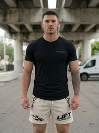 Man wearing black 'LIFT APPAREL' t-shirt and beige shorts standing under a bridge.