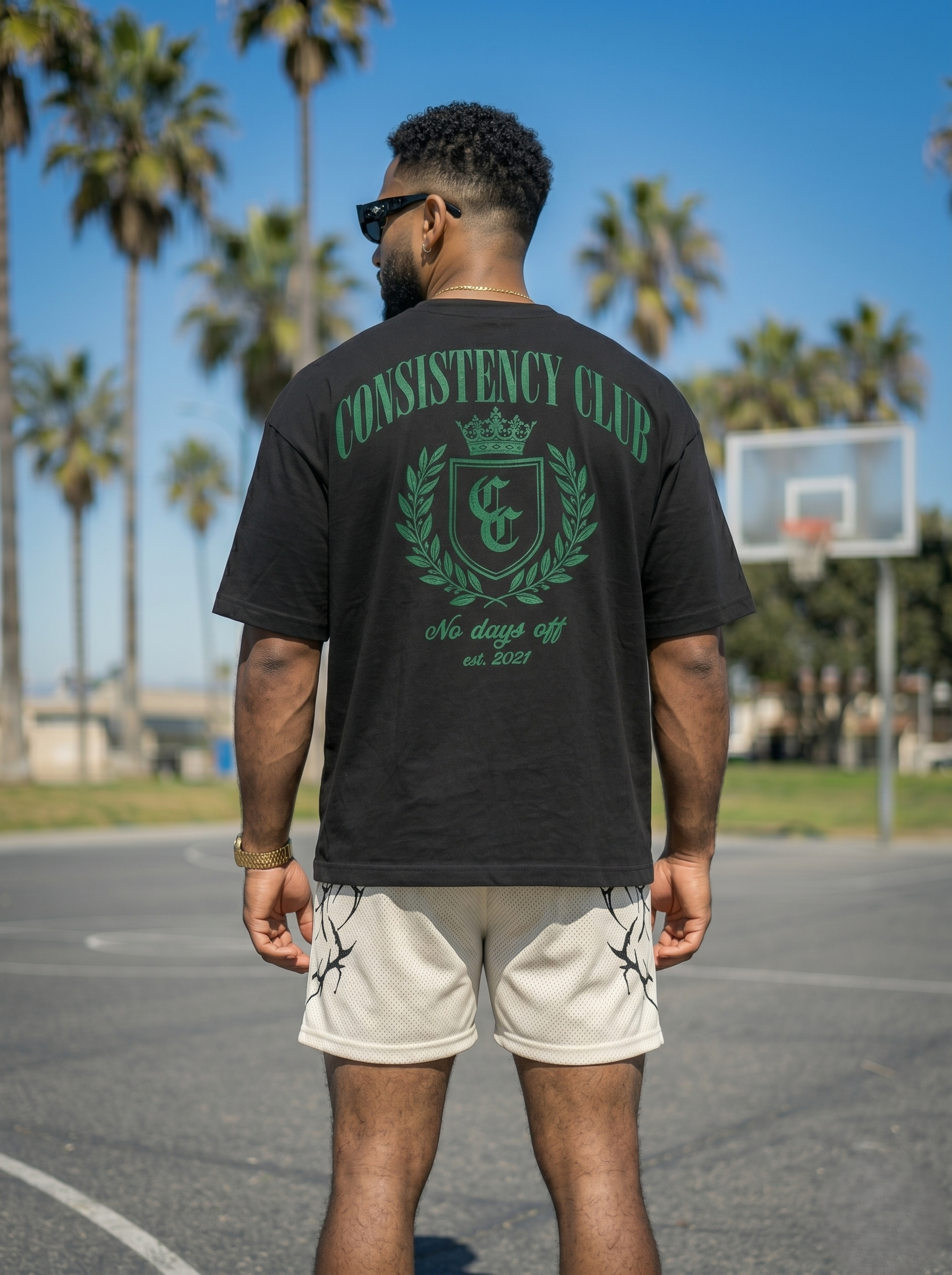 Man wearing a black 'Consistency Club' t-shirt on a basketball court with palm trees in the background