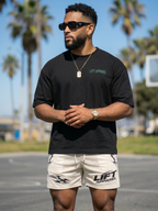 Man wearing a black 'LIFT APPAREL' t-shirt and beige shorts standing outdoors with palm trees in the background.
