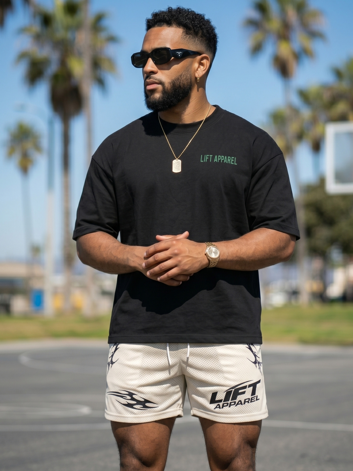 Man wearing a black 'LIFT APPAREL' t-shirt and beige shorts standing outdoors with palm trees in the background.