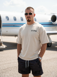 Man wearing sunglasses and a beige t-shirt with a logo, standing in front of a private jet.